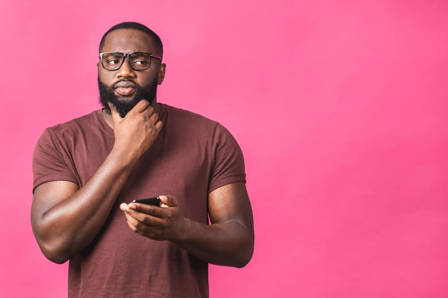 мислячий афроамериканець, одягнений у повсякденне надсилання та отримання повідомлень своїй коханій ізольовано на рожевому тлі. За допомогою телефону.