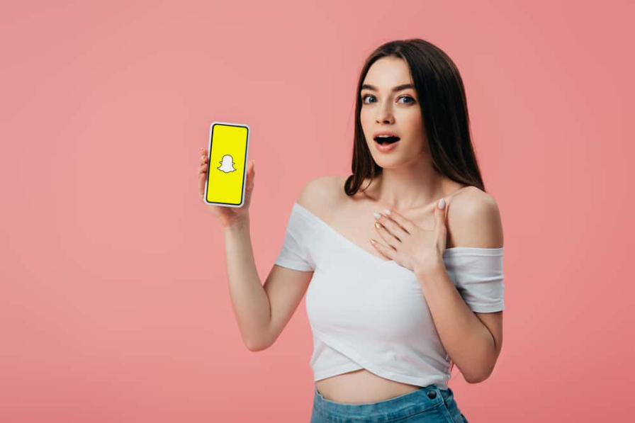 красива шокована дівчина тримає смартфон із додатком snapchat ізольовано на рожевому