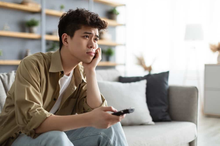 Нудна телевізійна програма. Нудно азіатський чоловік дивиться телевізор, перемикаючи канали за допомогою пульта дистанційного керування
