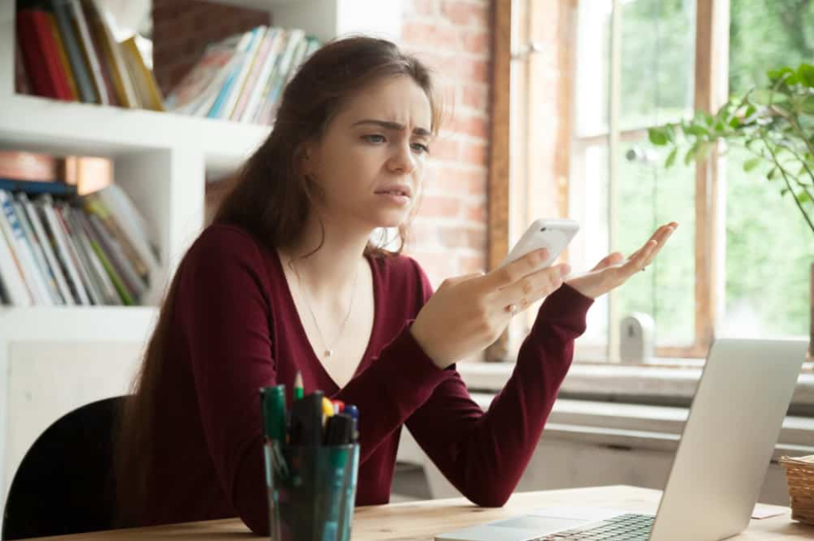 Розчарована жінка, яка має проблеми з непрацюючим смартфоном, сидячи за робочим столом вдома