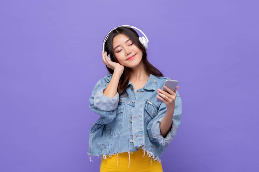 Щаслива азіатська дівчина в навушниках, слухаючи музику з мобільного телефону, відпочиває із закритими очима на фоні ізольованої фіолетової студії