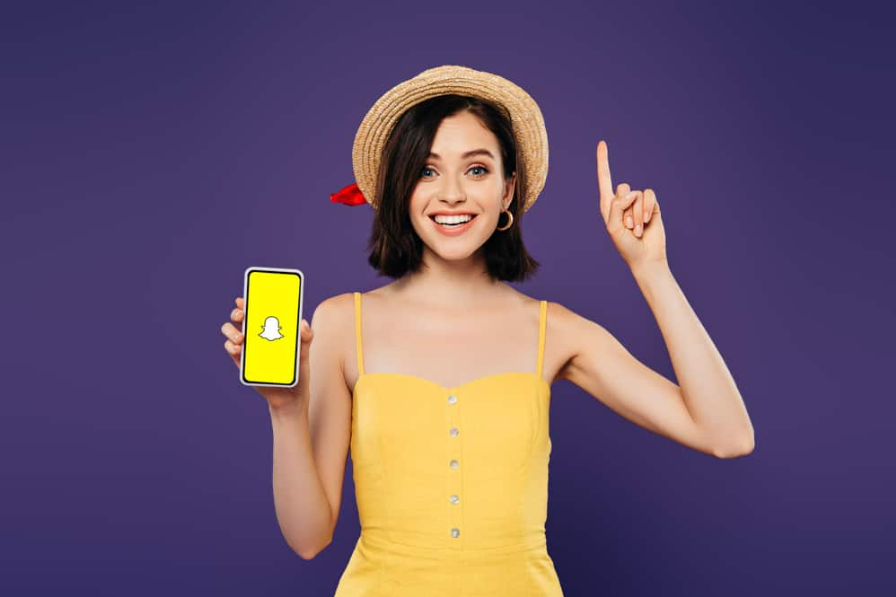 схвильована дівчина в солом'яному капелюсі показує ідею жесту та тримає смартфон із додатком snapchat, ізольованим на фіолетовому