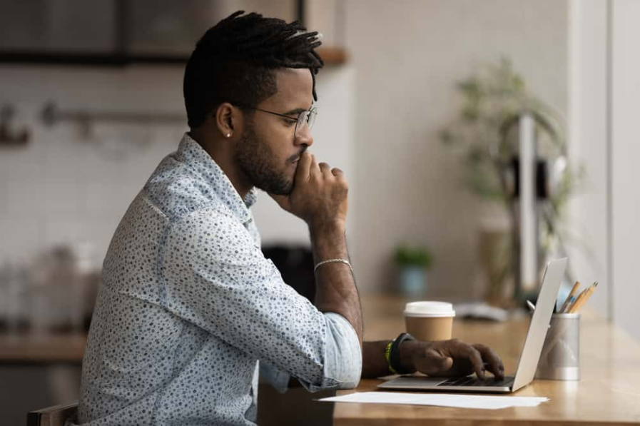 юнак двох расових категорій в окулярах сидить за столом і дивиться на екран ноутбука, працюючи онлайн в домашньому офісі