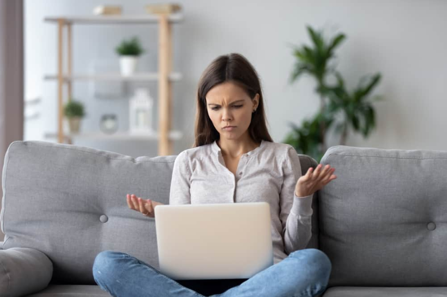 Збентежена молода жінка дивиться на ноутбук вдома, почуваючись збентеженою відсутністю зв’язку