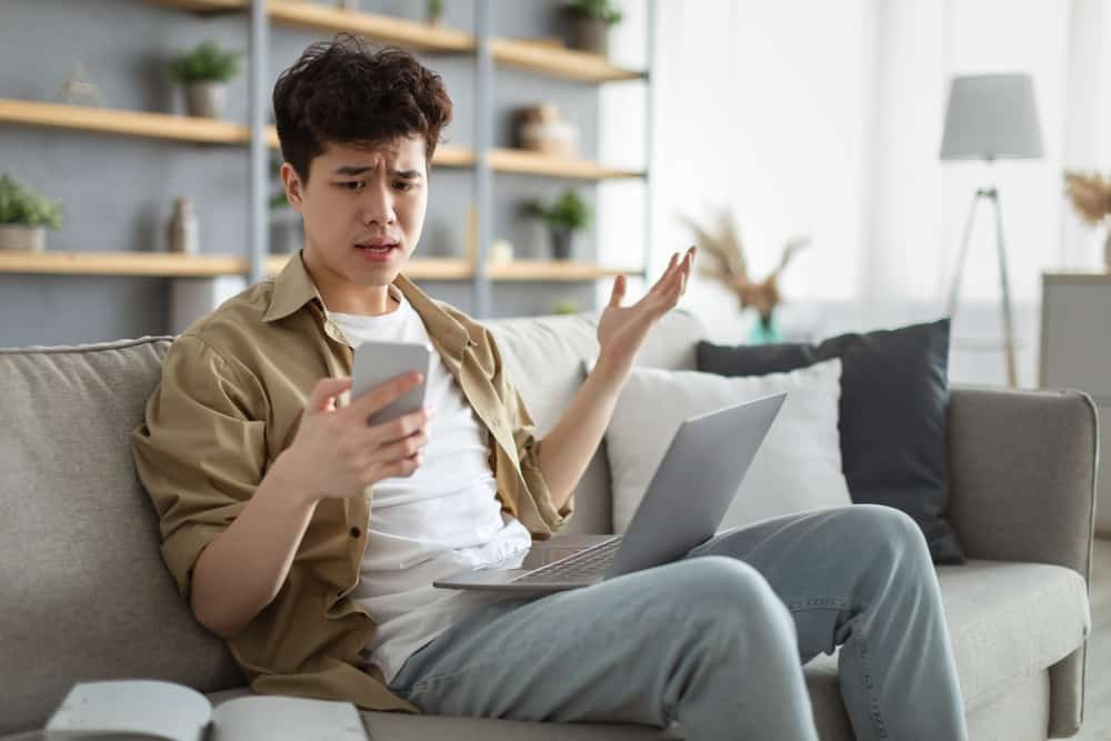 Розгублений азіатський чоловік дивиться на екран телефону, сидячи на дивані з комп’ютером на колінах у вітальні