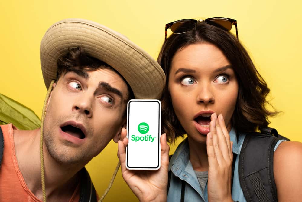 Здивована молода жінка показує смартфон із додатком Spotify, стоячи біля шокованого чоловіка на жовтому фоні.